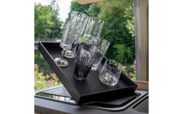 Schwarzes rechteckiges Tablett mit fünf Gläsern unterschiedlicher Form (zwei schlanke hohe Gläser, zwei mittelhohe Gläser, ein kurzes Glas) auf der Spülenkante; im Hintergrund Bäume, Wasser und rosa Blumen