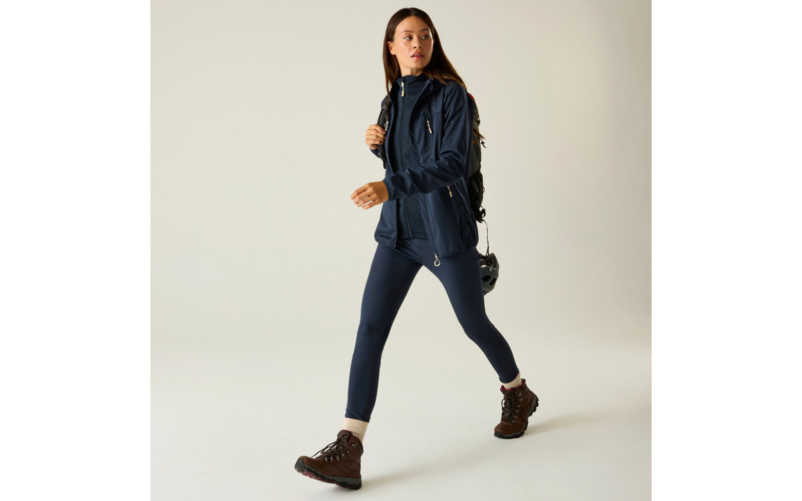 Mujer caminando hacia la derecha, lleva chaqueta y leggings azul marino, botas de montaña marrones, mochila oscura, fondo claro