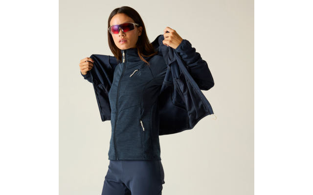 Persona que lleva una prenda superior ajustada y oscura con cremallera y pantalones oscuros, se está poniendo una chaqueta oscura, lleva gafas de sol espejadas, fondo liso y claro