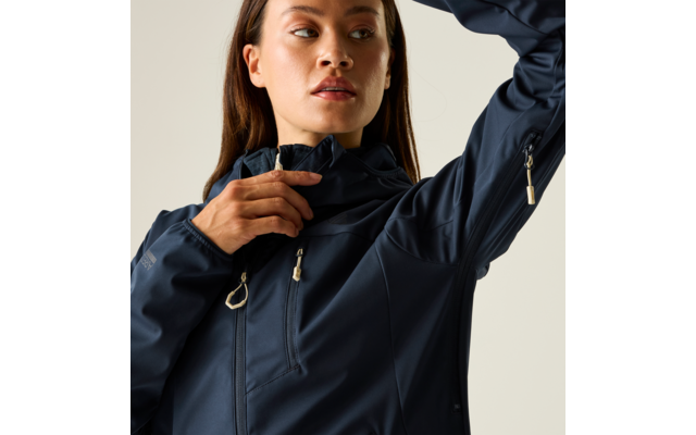 Persona con cabello castaño de longitud media que lleva una chaqueta azul oscuro con cremallera, mano junto a la cremallera del cuello, fondo liso claro