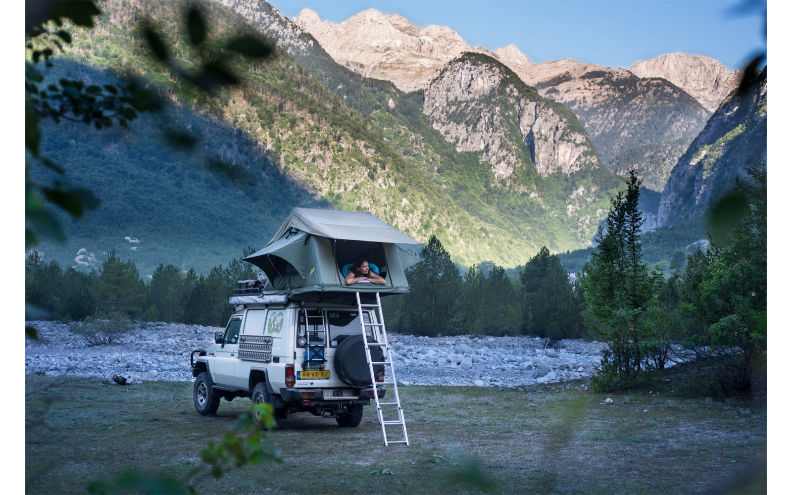 Geländewagen mit Dachzelt, aufgebaut neben einem Fluss und umgeben von Bergen und Wald