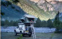 Geländewagen mit Dachzelt, aufgebaut neben einem Fluss und umgeben von Bergen und Wald