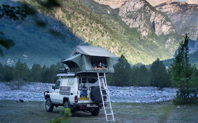 Geländewagen mit Dachzelt, aufgebaut neben einem Fluss und umgeben von Bergen und Wald