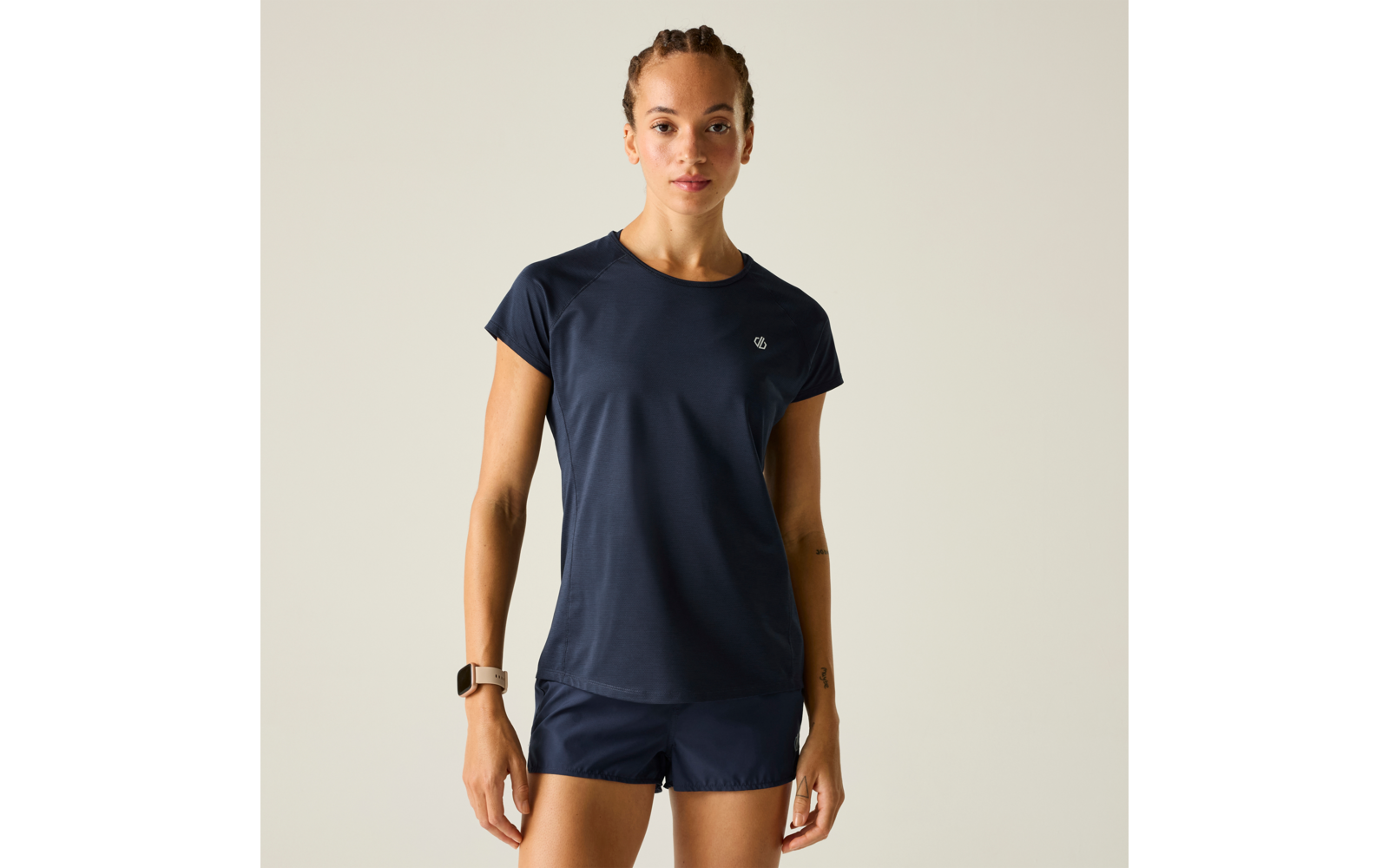 Frau steht frontal, trägt dunkelblaues T‑Shirt und dunkelblaue Shorts, Smartwatch am linken Handgelenk, einfarbiger heller Hintergrund