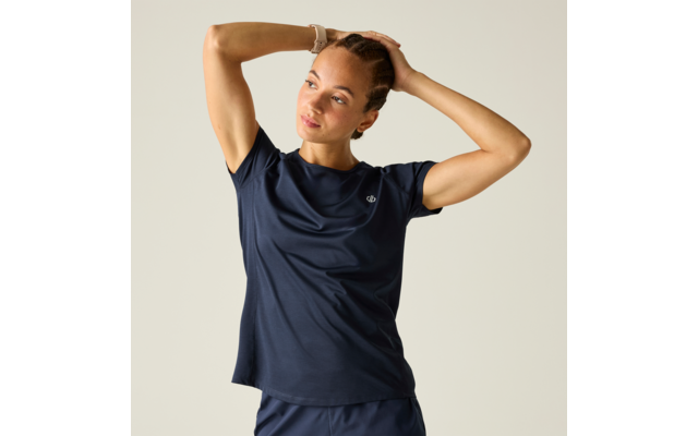 Person mit geflochtenen Haaren trägt dunkles blaues Sportshirt und Shorts, beide Hände hinter dem Kopf, Blick nach rechts, heller einfarbiger Hintergrund