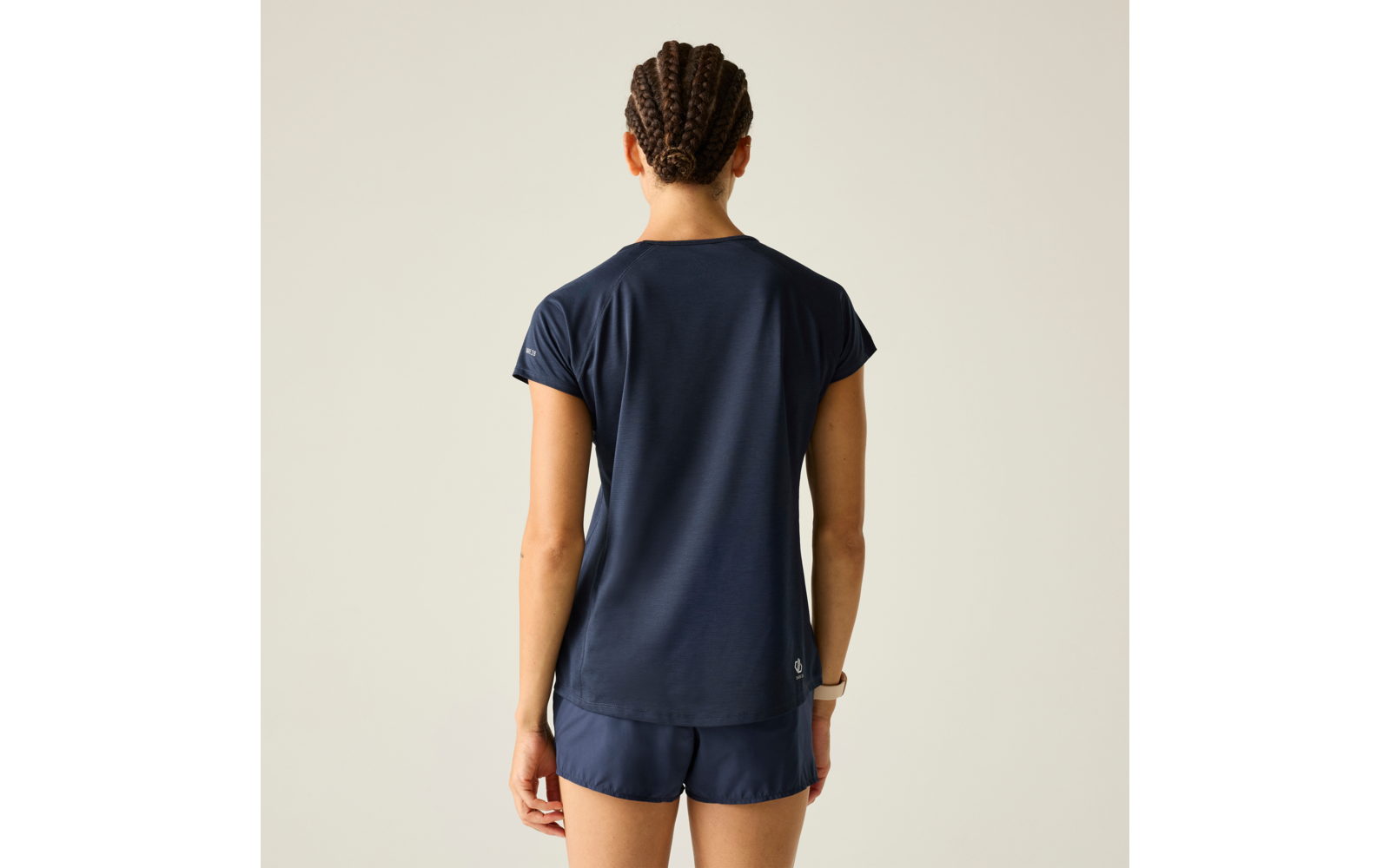 Person von hinten, dunkelblaues kurzärmeliges Shirt und kurze Sporthose, geflochtene Zöpfe, sichtbare Armbanduhr, heller einfarbiger Hintergrund