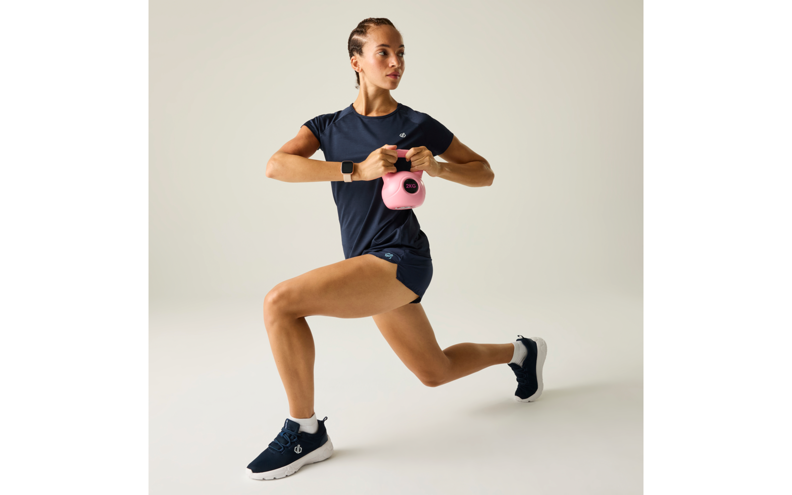 Frau in dunkelblauer Sportkleidung mit geflochtenem Haar hält eine pinke Kettlebell mit beiden Händen vor der Brust im Ausfallschritt, heller Hintergrund