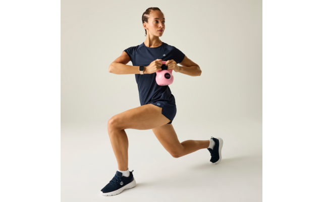 Frau in dunkelblauer Sportkleidung mit geflochtenem Haar hält eine pinke Kettlebell mit beiden Händen vor der Brust im Ausfallschritt, heller Hintergrund