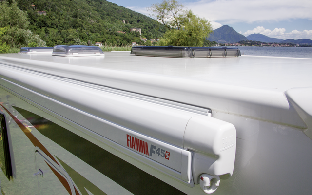 Dach eines weißen Wohnmobils mit zwei Dachlüftern und einer aufgerollten Markise mit Schriftzug „Fiamma F45S“, im Hintergrund See, Bäume und Berge unter blauem Himmel