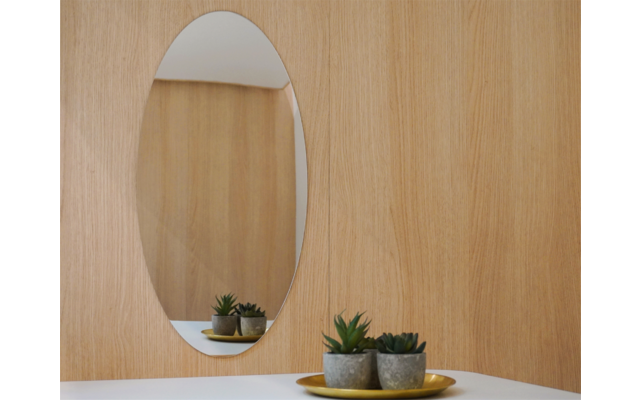 Ovaler Wandspiegel über weißer Ablage; drei kleine Sukkulenten in grauen Töpfen auf einem goldfarbenen Tablett auf der Ablage; Spiegel spiegelt eine helle Holzwand