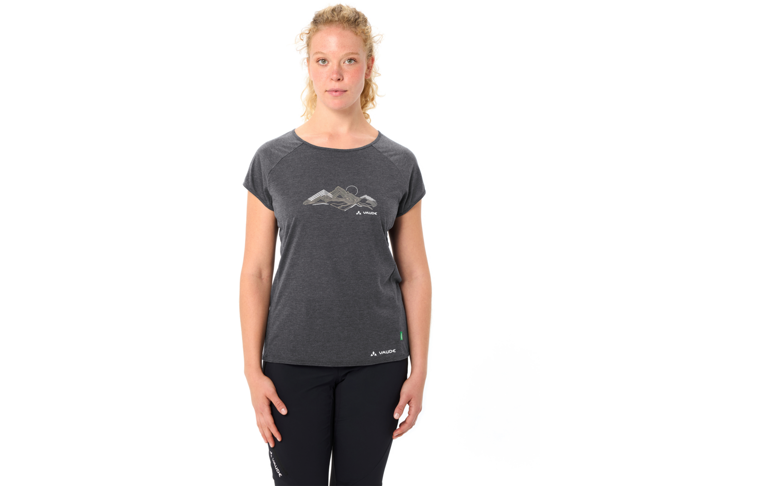 Person steht frontal vor weißem Hintergrund, trägt dunkelgraues T‑Shirt mit Bergmotiv und schwarze Hose, Haare zurückgebunden