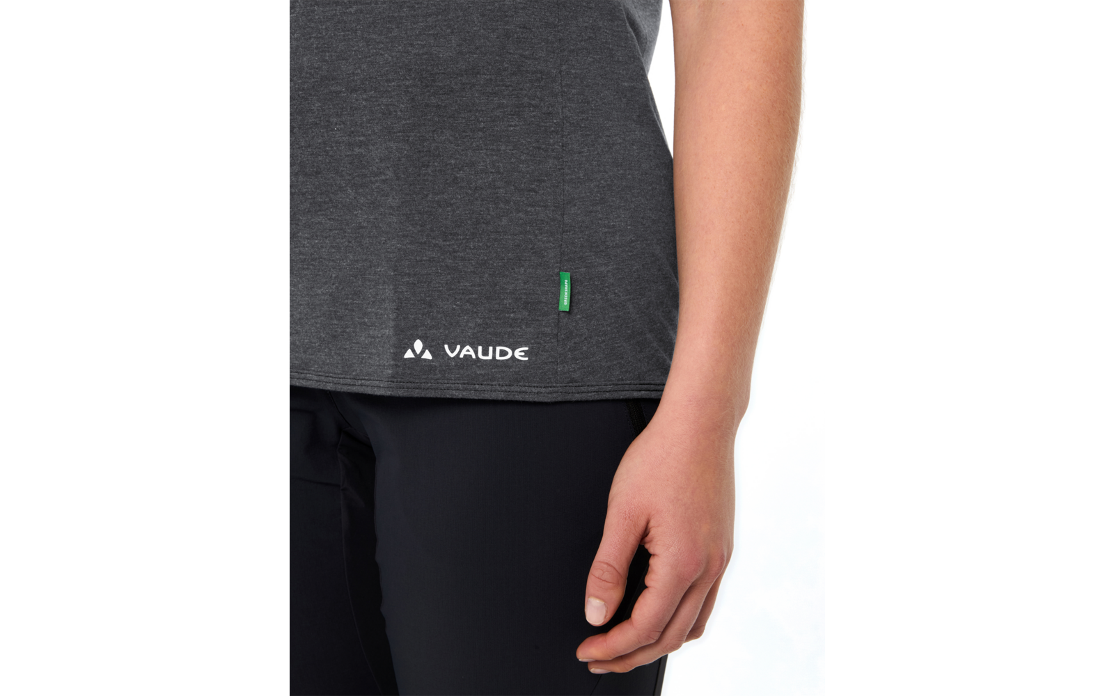 Unterer Teil eines dunkelgrauen T-Shirts mit VAUDE-Logo und kleinem grünem Etikett, schwarze Hose, rechte Hand an der Seite