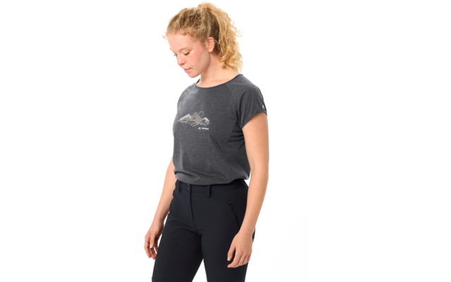 Person mit blondem, lockigem Pferdeschwanz, schaut nach unten, trägt dunkelgraues T‑Shirt mit kleinem Bergmotiv und dunkle Hose vor weißem Hintergrund