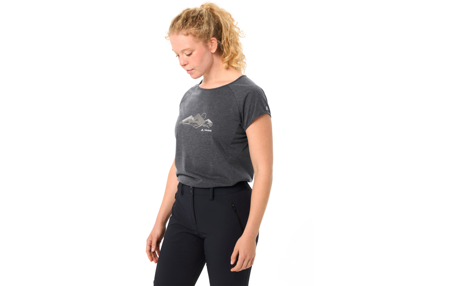 Person mit blondem, lockigem Pferdeschwanz, schaut nach unten, trägt dunkelgraues T‑Shirt mit kleinem Bergmotiv und dunkle Hose vor weißem Hintergrund