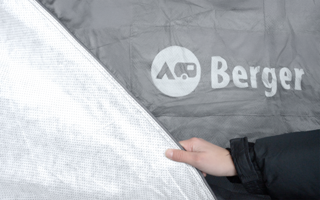 Mano con manga de chaqueta oscura que aparta una tela plateada con puntos; detrás, una superficie de tela gris con el texto blanco Berger y un símbolo circular