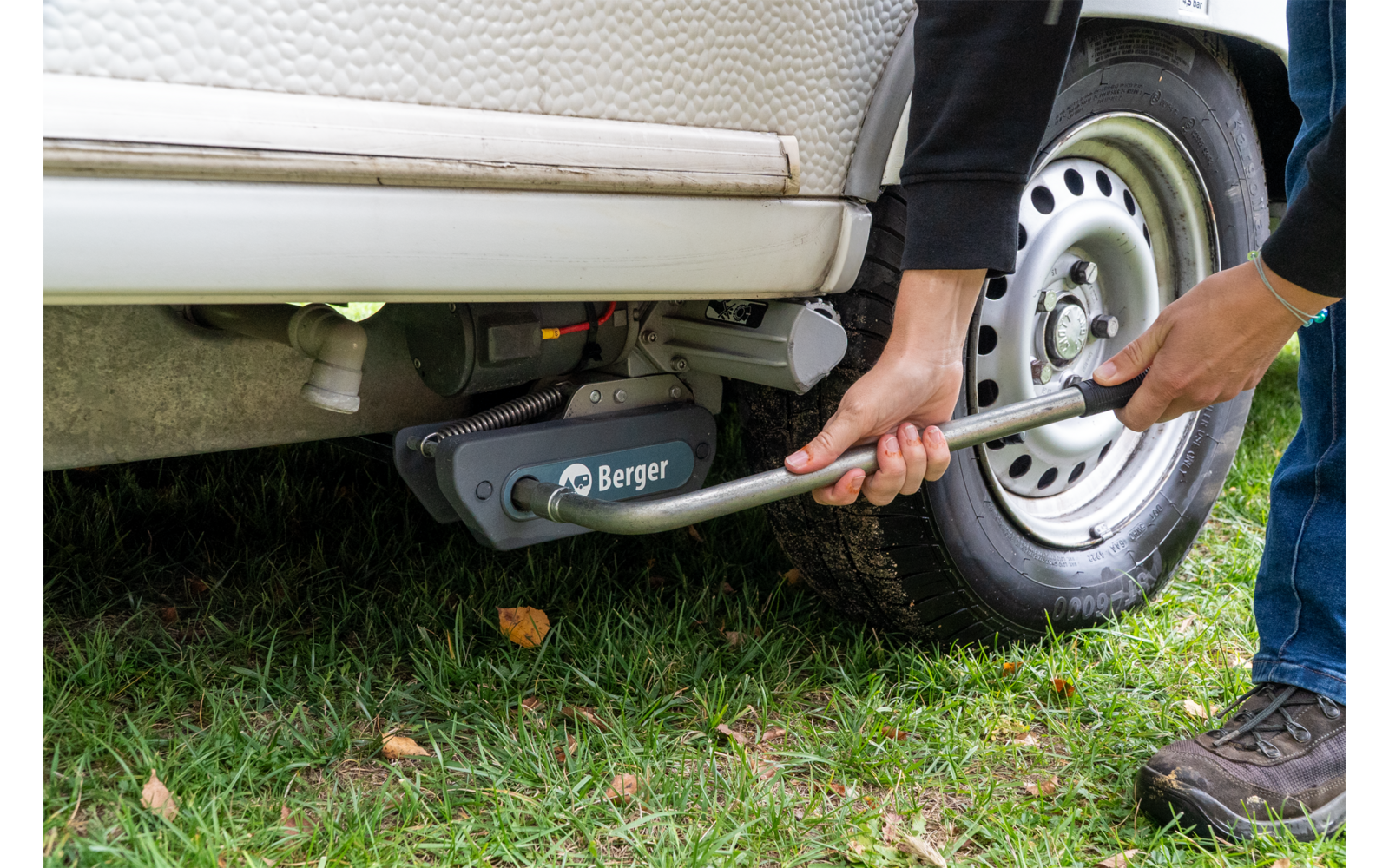 Hände drehen die Kurbel eines Wagenhebers unter einem Anhänger neben einem Rad; Person trägt Jeans und Stiefel; Grasboden