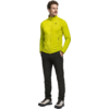 Ganzkörperaufnahme einer Person in gelber Fleecejacke mit Reißverschluss und kleinem Logo, schwarzen Hosen und weißen Sneakers, Hände in den Taschen, schaut nach rechts