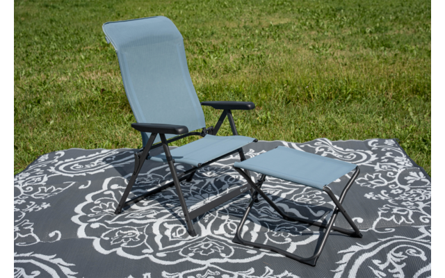 Hellblauer Klappstuhl mit schwarzem Metallgestell und passendem Fußhocker auf grauem Teppich mit weißem Blumenmuster auf Gras