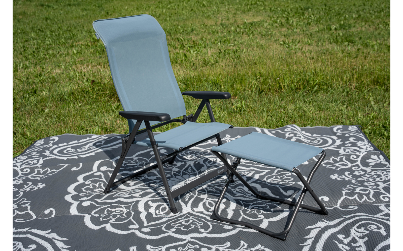Hellblauer Klappstuhl mit schwarzem Metallgestell und passendem Fußhocker auf grauem Teppich mit weißem Blumenmuster auf Gras