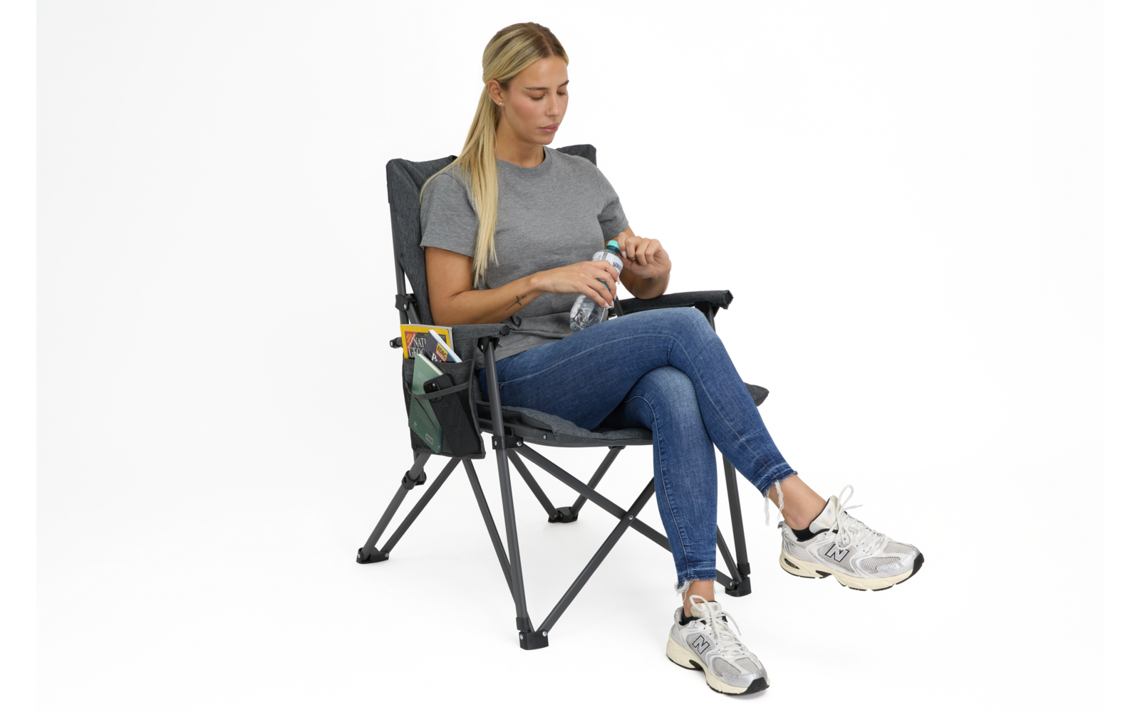 Person sitzt in faltbarem Campingstuhl, hält eine Wasserflasche, trägt graues T-Shirt, blaue Jeans und weiße Sneakers