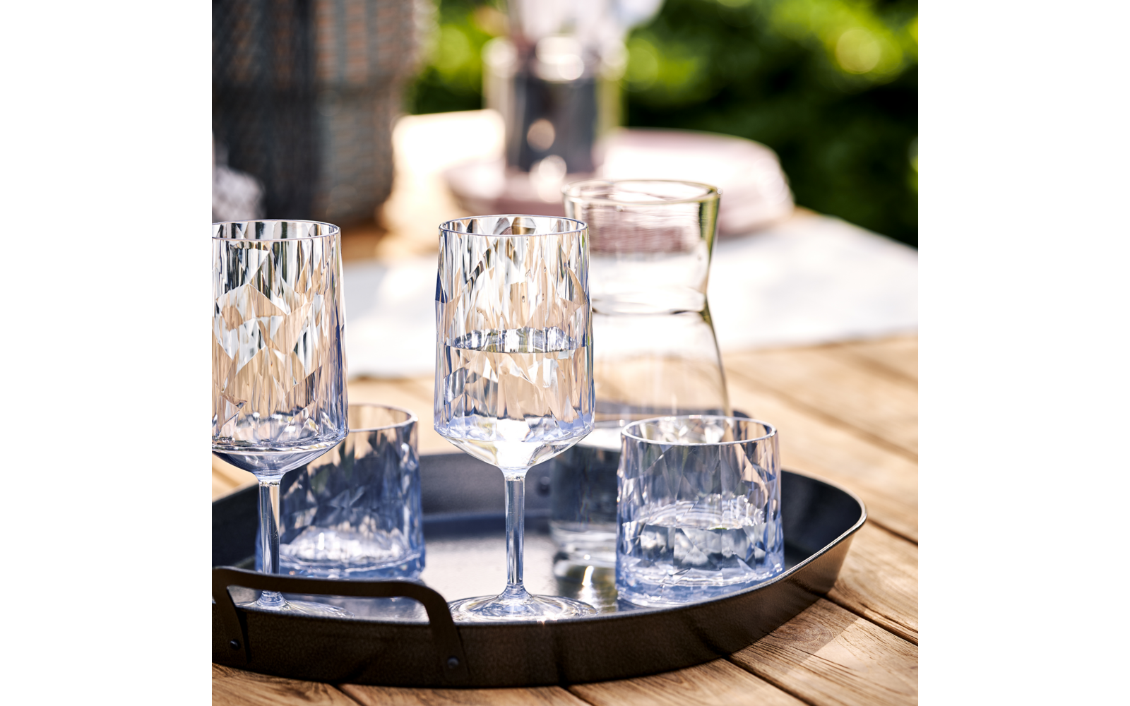 Schwarzes Tablett mit zwei hohen Stielgläsern, zwei niedrigen Tumblern und einer klaren Glas-Karaffe auf einem Holztisch, unscharfer grüner Hintergrund