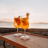 Zwei Weingläser mit orangefarbenem Cocktail, jeweils garniert mit einer Orangenscheibe und einem Ananasblatt, auf einem Holztablett vor Strand und Meer