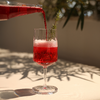 Rote Flüssigkeit, die aus einer Flasche in ein Stielglas mit Schaum gegossen wird; Rosmarinzweig im Glas, auf hellem Tisch mit Schatten und unscharfem Pflanzenhintergrund