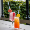 Zwei Gläser mit farbigen Getränken auf einem Tisch; vorn ein hohes Glas mit orange-rotem Getränk, Eis und gelber Zitrusscheibe am Rand, hinten ein Weinglas mit rosa Getränk, Eis und roten Fruchtstücken; beide mit Strohhalmen, unscharfer grüner Hinter