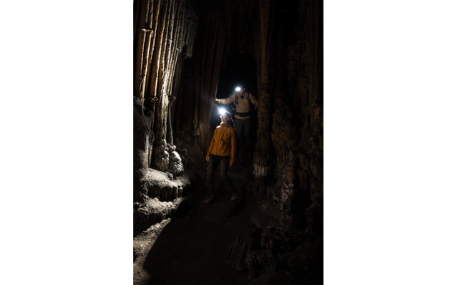 Deux personnes avec lampes frontales dans un passage de grotte sombre avec stalactites et stalagmites ; personne de devant vêtue d'une veste jaune, personne derrière touche la paroi rocheuse