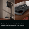 Split image: left a stick vacuum on a wall mount next to an outlet with visible cable; right a close-up of the handheld unit with transparent dust container on a wooden surface; black bar with German text at bottom