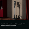 Stick vacuum in a freestanding dock beside a wall outlet, two accessories on the floor, red cabinet left, red curtain right, wooden floor, gray wall