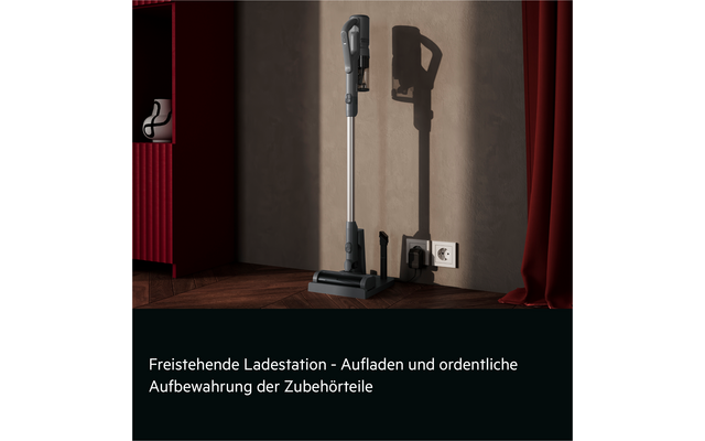 Stick vacuum in a freestanding dock beside a wall outlet, two accessories on the floor, red cabinet left, red curtain right, wooden floor, gray wall
