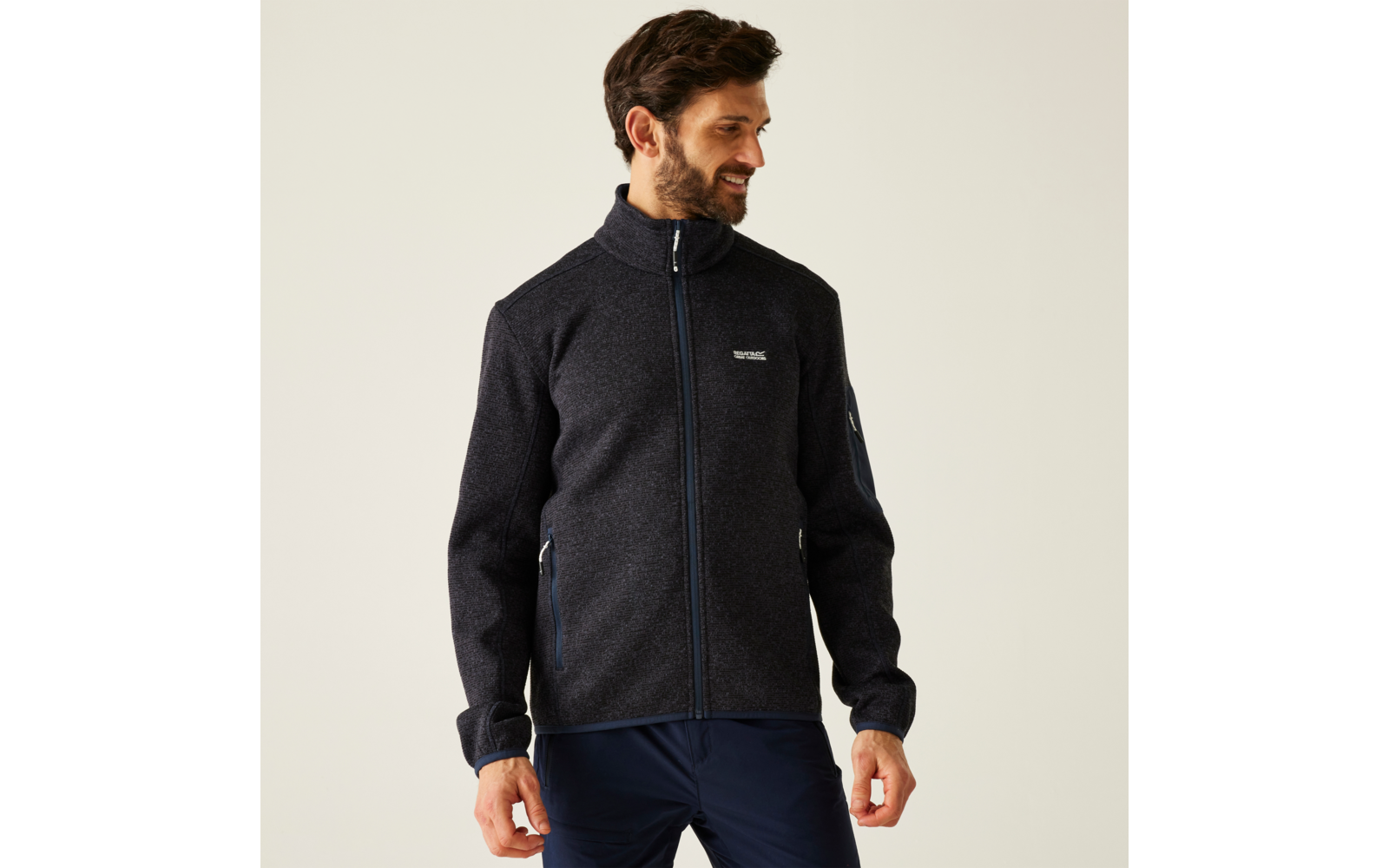 Man met baard draagt donkergrijze fleecejas met rits en blauwe broek, staat voor een lichte achtergrond en kijkt opzij