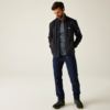 Man met donker haar kijkt omlaag, draagt donkergrijs fleecejack, grijs T‑shirt, donkerblauwe broek, zwarte wandelschoenen met groene accenten, handen in de zakken, effen lichte achtergrond