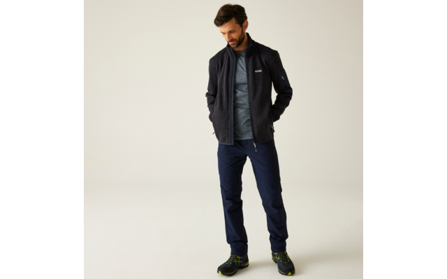 Man met donker haar kijkt omlaag, draagt donkergrijs fleecejack, grijs T‑shirt, donkerblauwe broek, zwarte wandelschoenen met groene accenten, handen in de zakken, effen lichte achtergrond