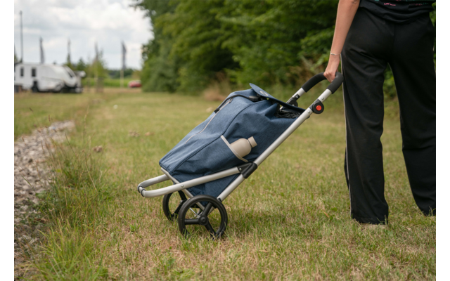 Lower body of a person in black pants, hand on the handle of a blue shopping trolley, trolley on grass beside a gravel path, trees and white vehicles in the background