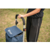 Person shown from the waist down, right hand on the handle of a blue shopping trolley with visible logo, grass in the background