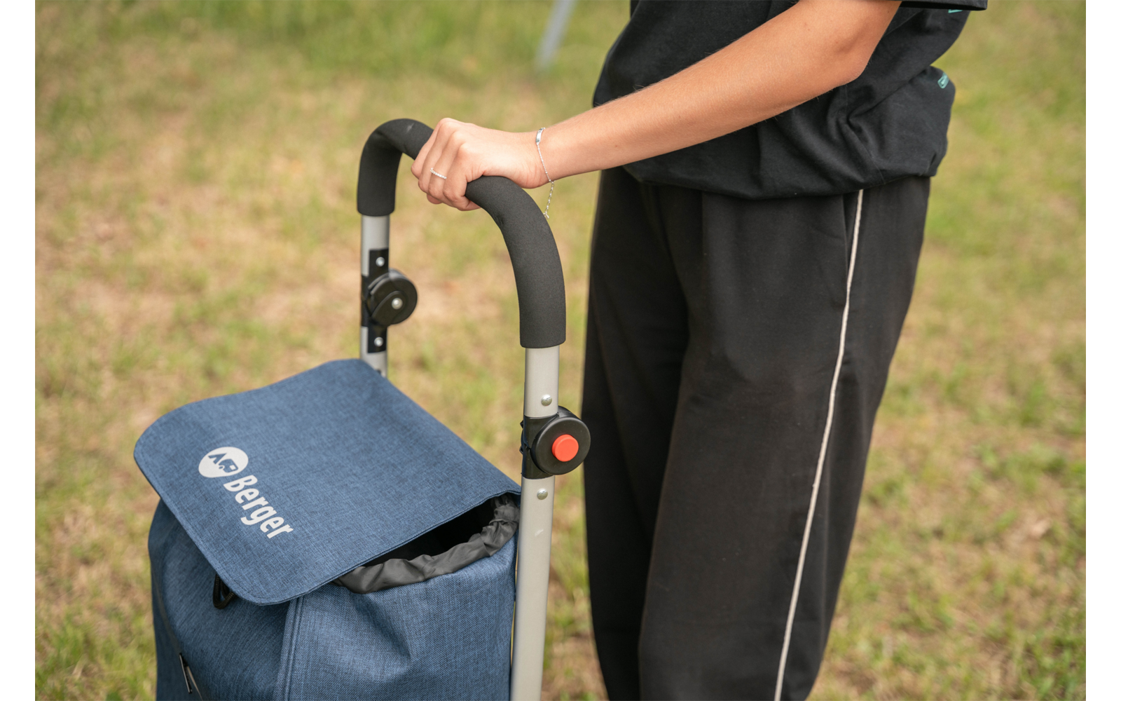 Person shown from the waist down, right hand on the handle of a blue shopping trolley with visible logo, grass in the background