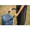 Two hands holding the black handle of a blue shopping trolley with a flap; person wearing black pants; grass in the background
