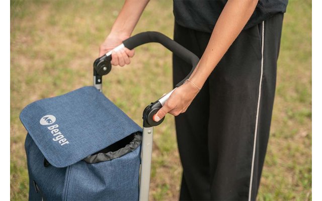 Two hands holding the black handle of a blue shopping trolley with a flap; person wearing black pants; grass in the background