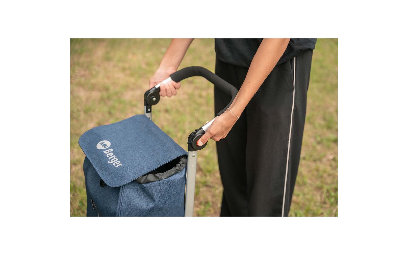 Two hands holding the black handle of a blue shopping trolley with a flap; person wearing black pants; grass in the background