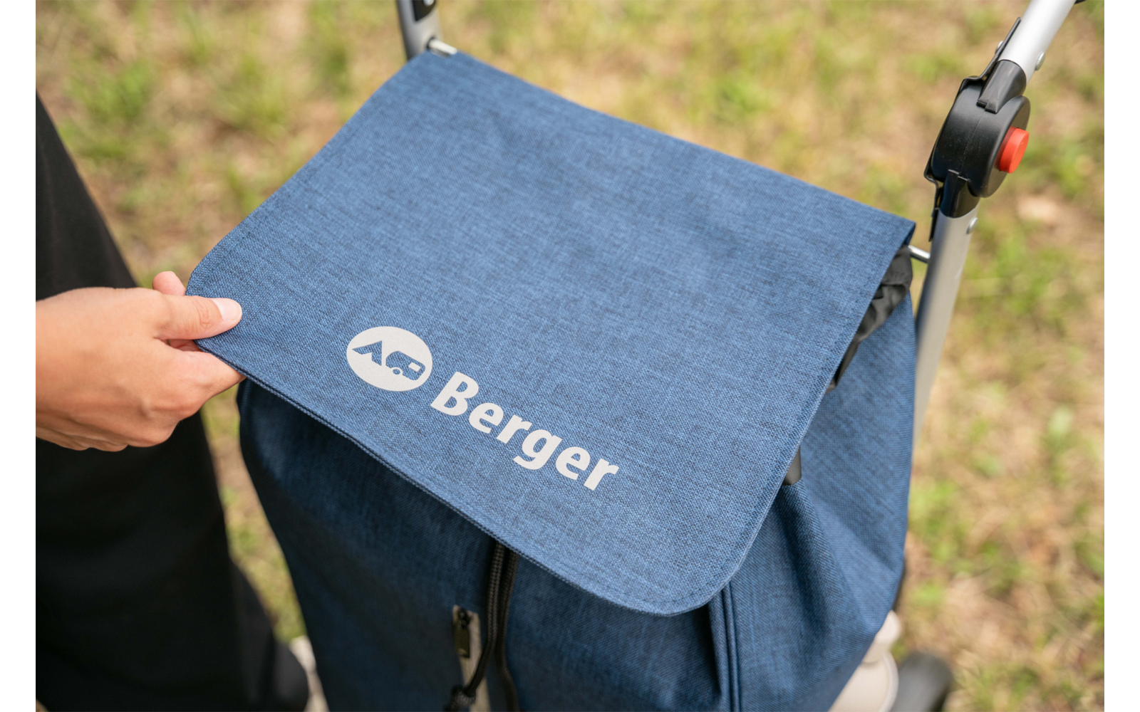Hand holding the flap of a blue bag with white 'AG Berger' logo attached to a stroller; grass in the background