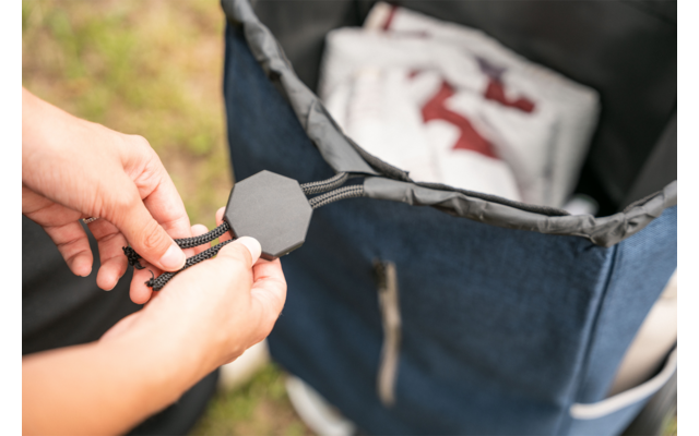 Two hands pulling a black cord lock at the opening of a blue backpack; a white garment with a red pattern is visible inside the backpack