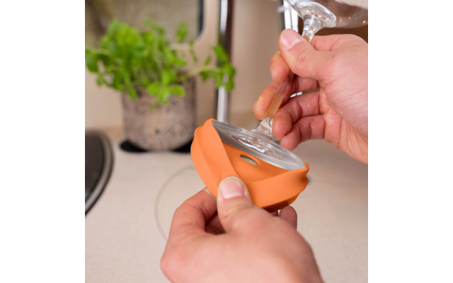 Handen plaatsen een oranje siliconen hoes rond de voet van een wijnglas op een keukenaanrecht; plant en gootsteen op de achtergrond