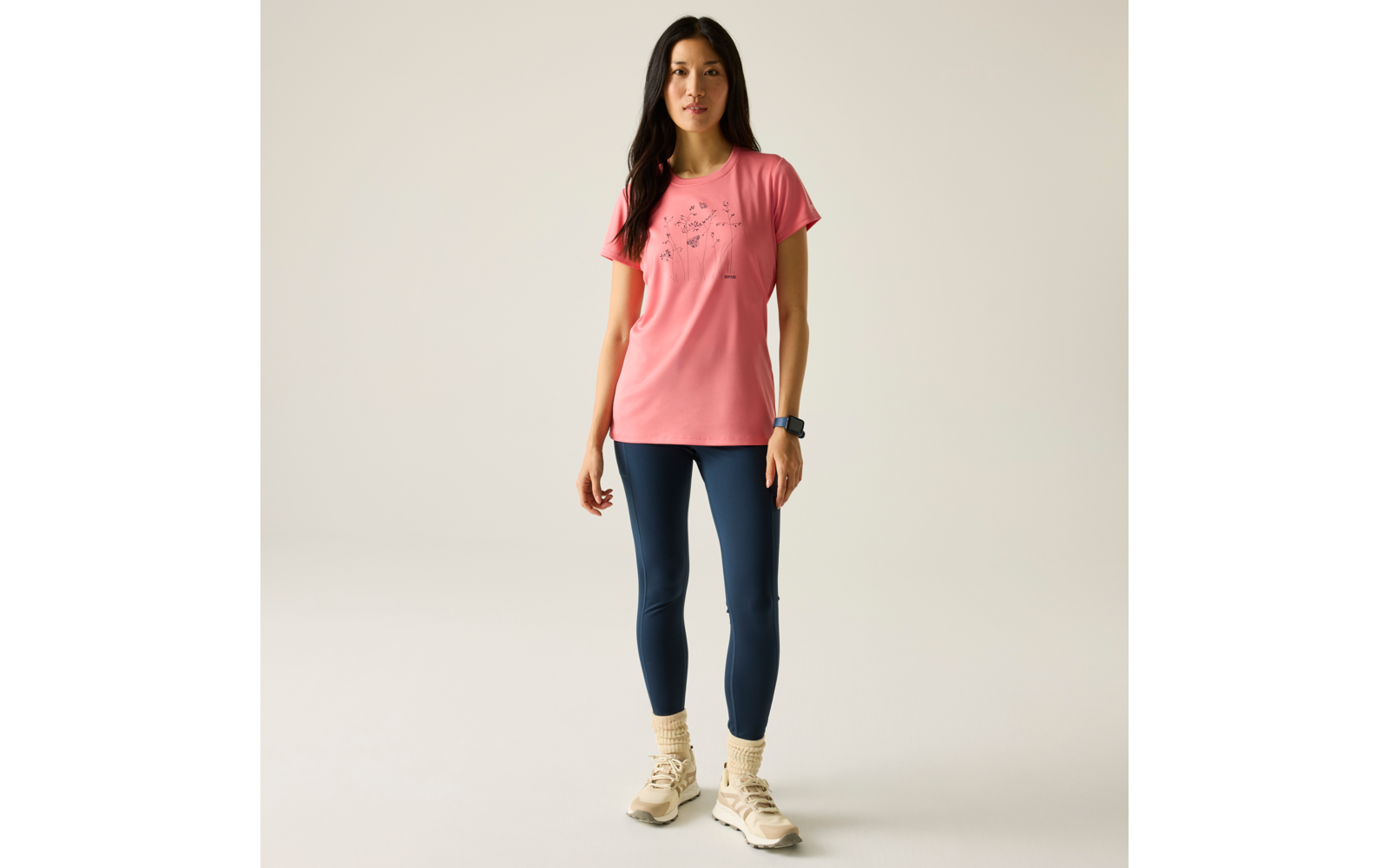 Personne debout devant un fond clair, portant un t‑shirt rose avec petit motif, leggings bleu marine, baskets claires, chaussettes en maille et une montre au poignet gauche