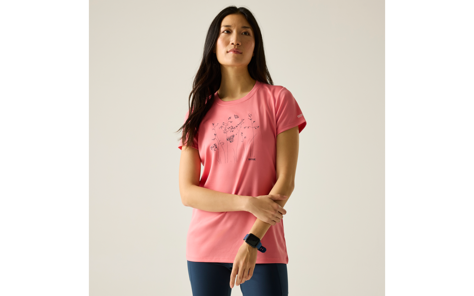 Femme debout devant un fond clair, portant un t‑shirt rose à motif floral, un legging bleu foncé et une montre connectée au poignet gauche