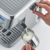 Hand dreht Drehknopf an Espressomaschine; Dampfdüse im Edelstahl-Milchkännchen mit sichtbarem Milchschaum