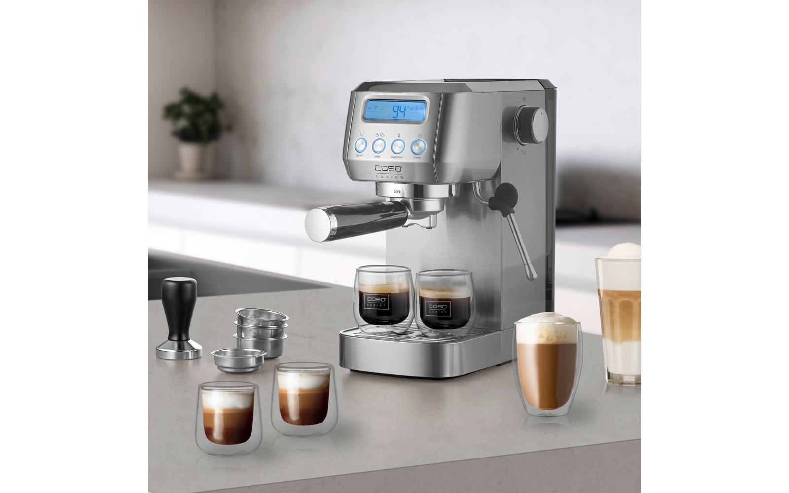 Edelstahl-Espressomaschine auf Küchenarbeitsplatte, zwei Espressotassen unter dem Siebträger, weitere geschichtete Kaffees in Gläsern, Tamper, Filtereinsätze und Dampfdüse sichtbar