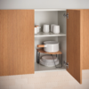 Offener Holzschrank mit zwei Türen und zwei Regalfächern. Oben drei weiße Tassen und zwei kleine weiße Gläser/Dosen. Unten ein weißer Topf mit Holzgriff auf einem kleinen Holzbrett mit schwarzen Metallbeinen, darunter ein Stapel weißer Teller