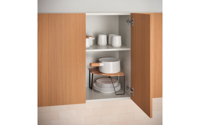 Offener Holzschrank mit zwei Türen und zwei Regalfächern. Oben drei weiße Tassen und zwei kleine weiße Gläser/Dosen. Unten ein weißer Topf mit Holzgriff auf einem kleinen Holzbrett mit schwarzen Metallbeinen, darunter ein Stapel weißer Teller